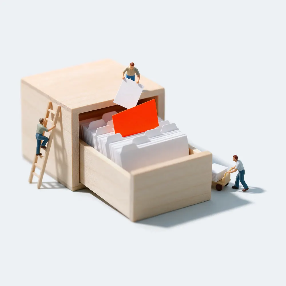 Ein offener Karteikasten voller weißer Karteikarten, eine Karte ist orange. Miniaturfiguren sortieren Karten ein.